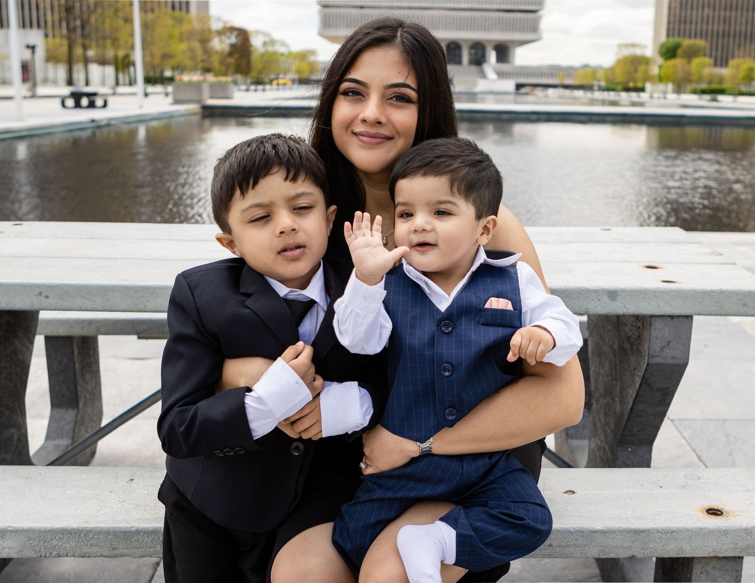 A smiling mother holds two young boys in dressy outfits at a picnic table beside a calm reflecting pool.
