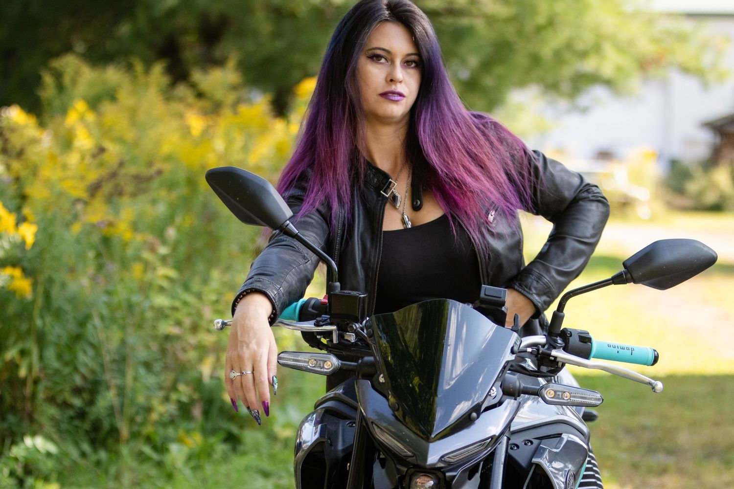 Woman with long purple hair and a black leather jacket sits on a black motorcycle beside yellow wildflowers.