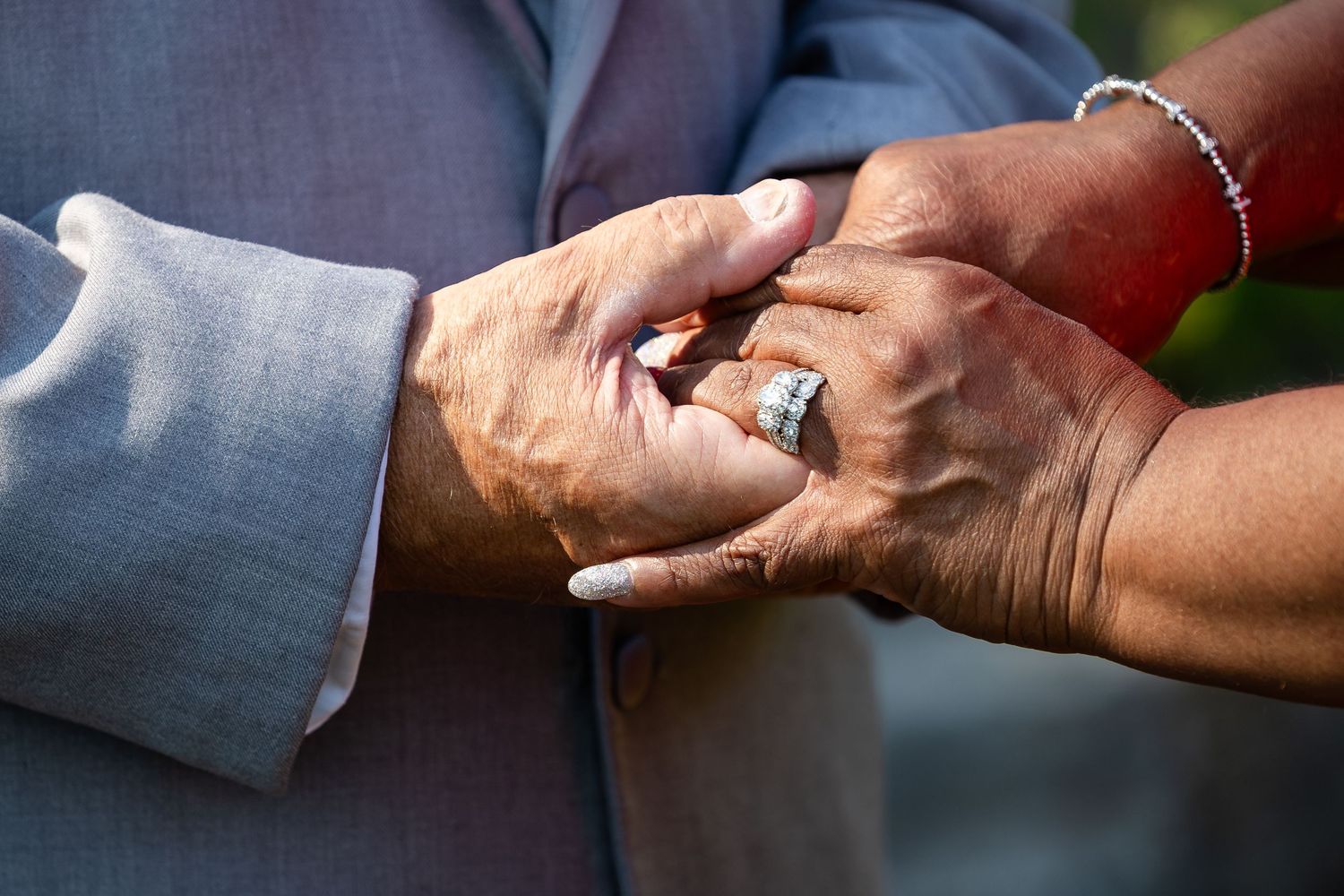 Close-up of an older couple's clasped hands, with a diamond wedding ring and beaded bracelet visible against a gray suit sleeve.