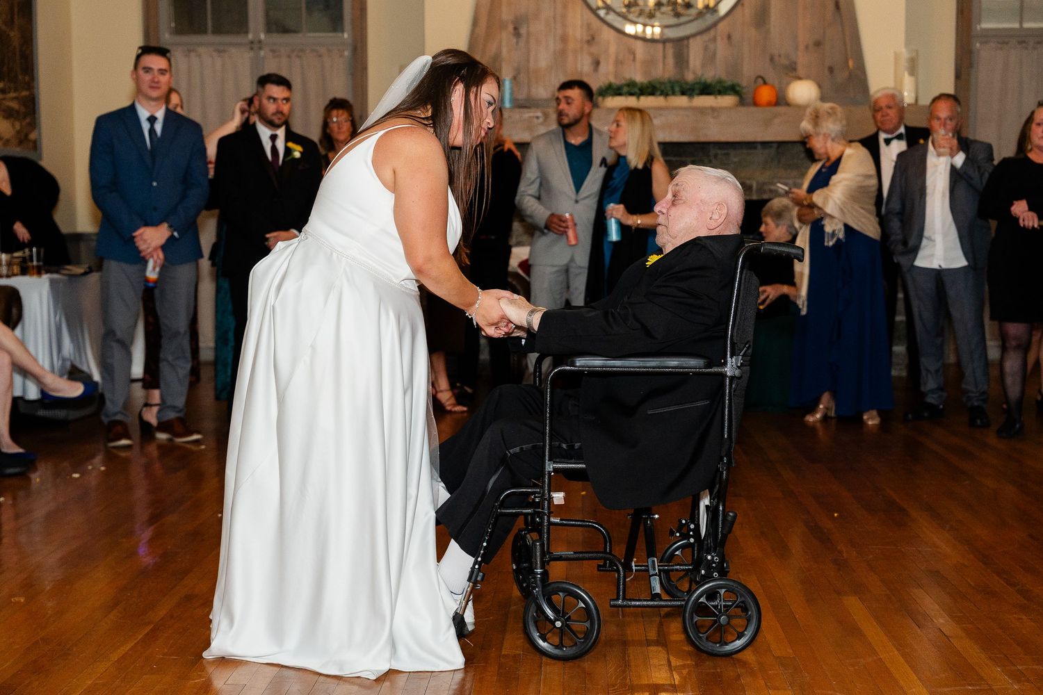 Bride in a white gown holds the hand of an elderly man in a wheelchair as wedding guests look on in a rustic reception hall.