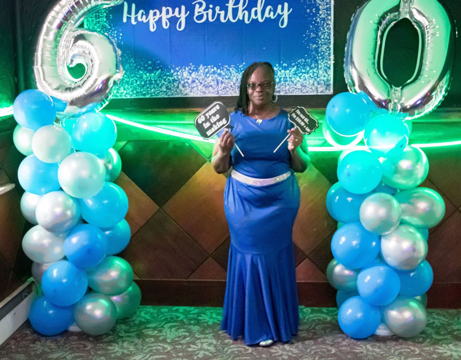 Woman in a blue gown holding photo props poses between blue and silver balloon columns with 6 and 0 numbers under a Happy Birthday banner.