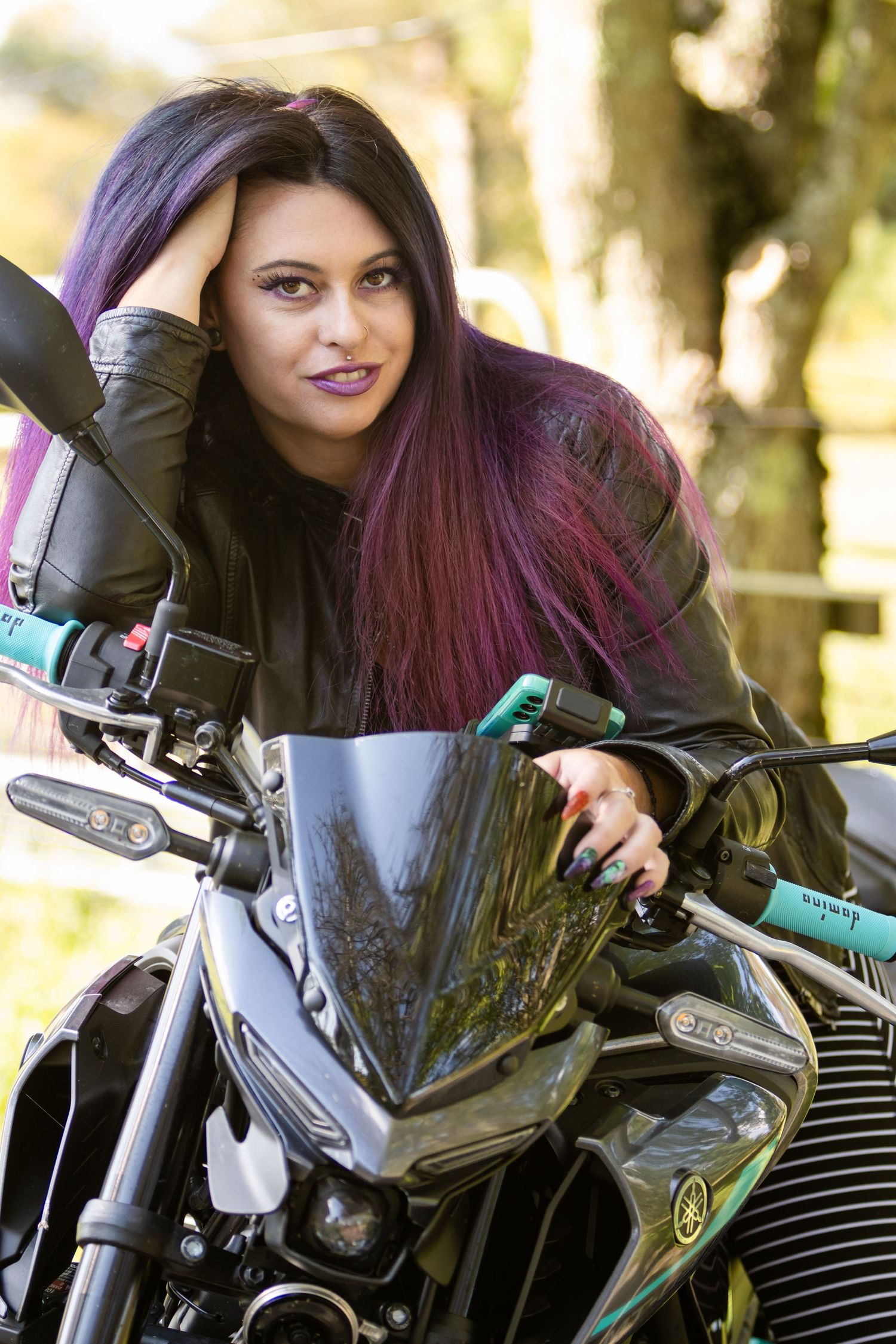 Woman with long purple ombre hair and black leather jacket leans on a sport motorcycle handlebar and smiles outdoors.