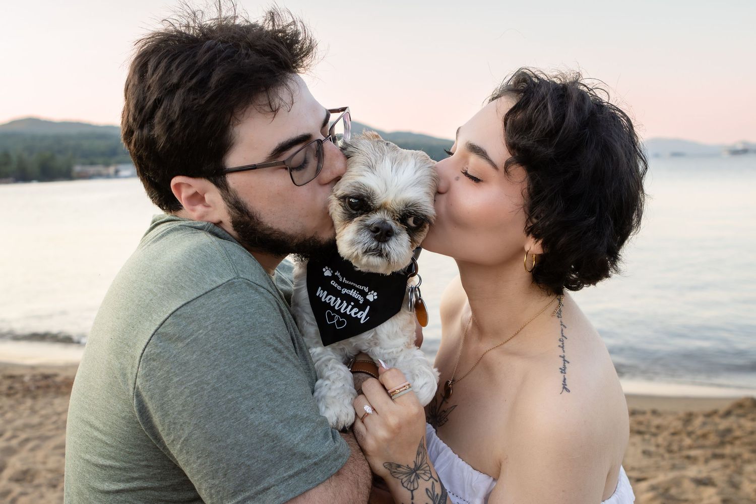 Couple kisses a shih tzu wearing a bandana on a lakeside beach at sunset.