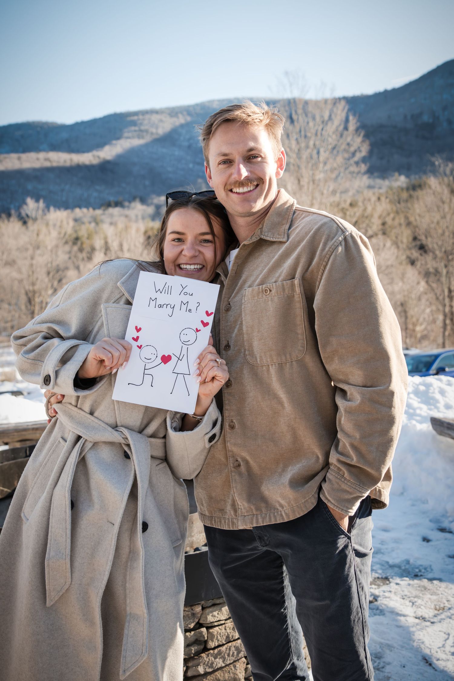 A smiling couple stands outdoors in winter as the woman holds a handmade card reading Will You Marry Me with a mountain backdrop.