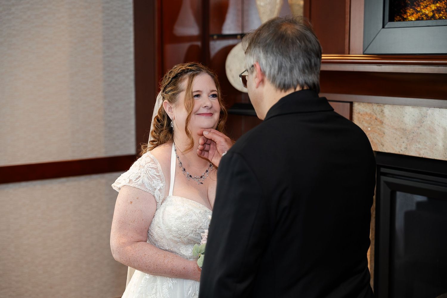 A smiling bride in a white lace wedding gown looks up at an older man in a black jacket during an indoor ceremony.