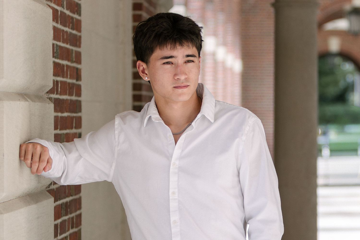 Young man in a white button-up shirt leans against a brick column in an outdoor covered walkway.