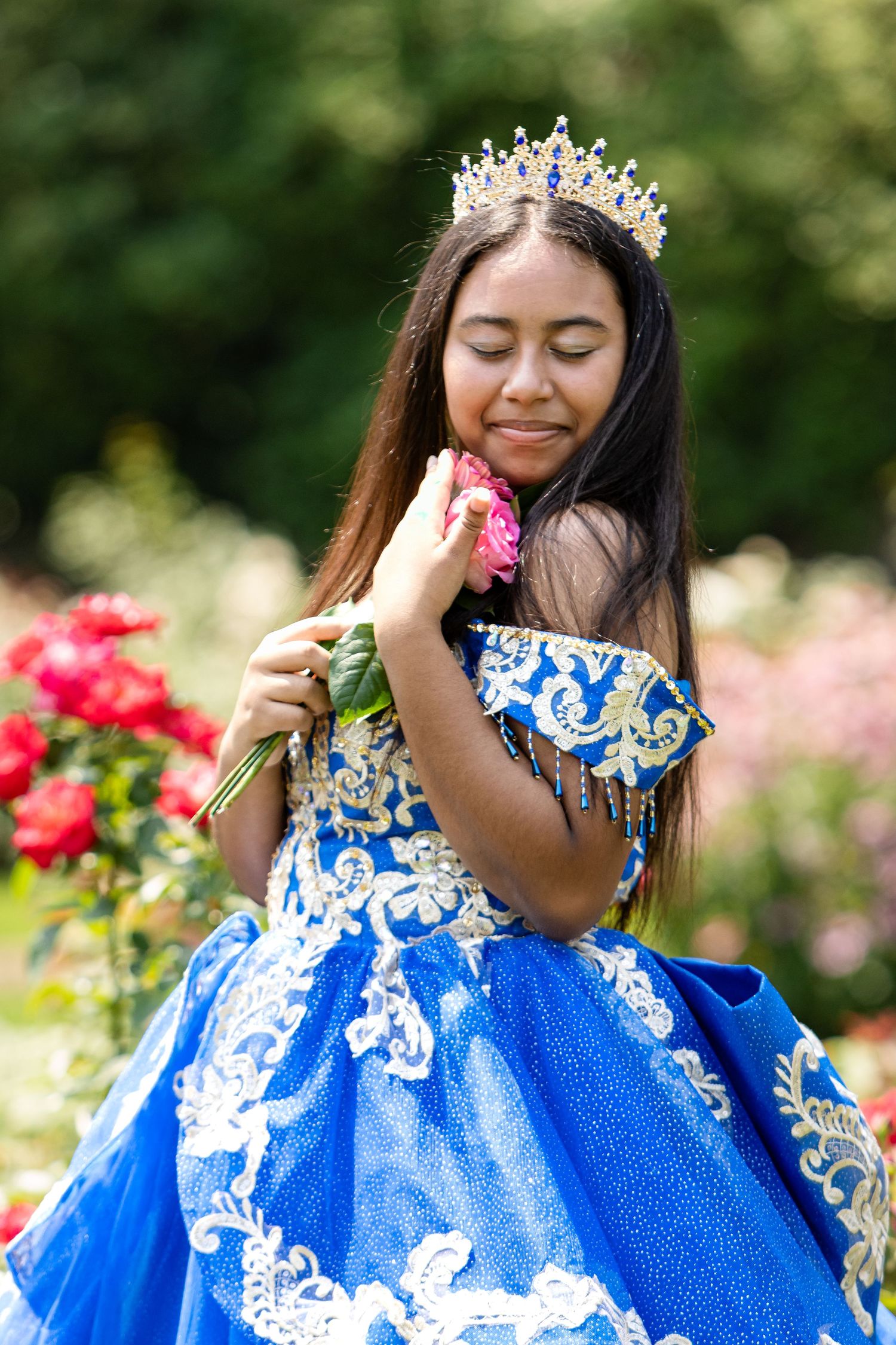 Girl in a sparkling blue embroidered quinceañera gown and tiara smells a pink rose in a lush garden.