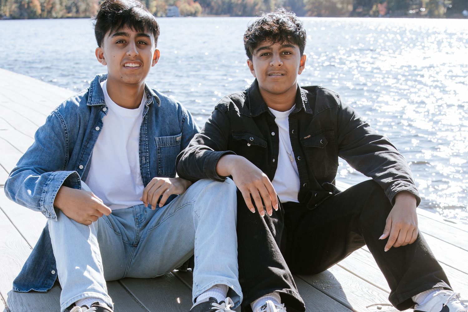 Two smiling young men in denim jackets sit side by side on a wooden dock beside a sparkling lake on a sunny day.