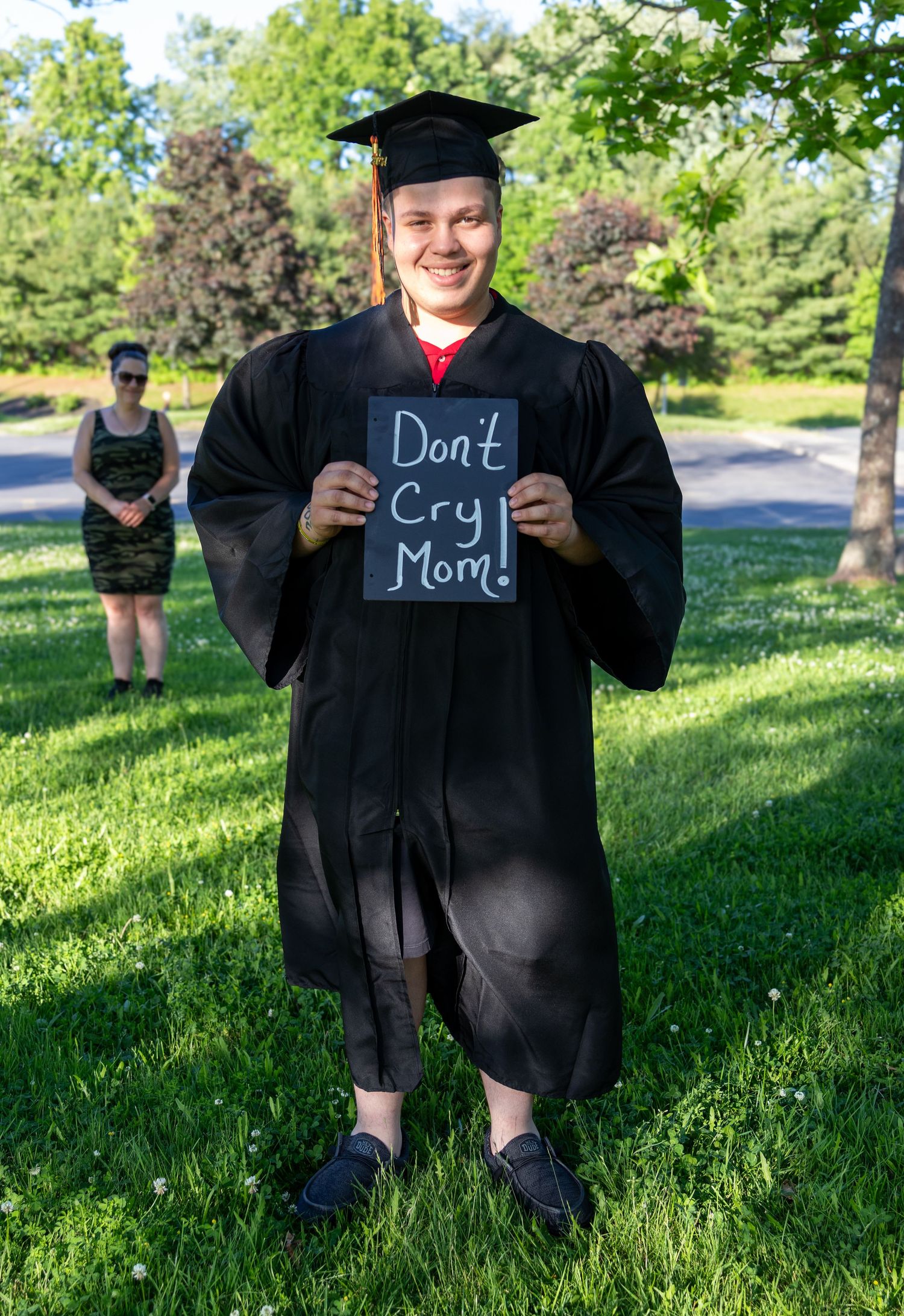 A smiling graduate in a black cap and gown holds a chalkboard sign reading Don't Cry Mom while a woman stands on the grass behind.