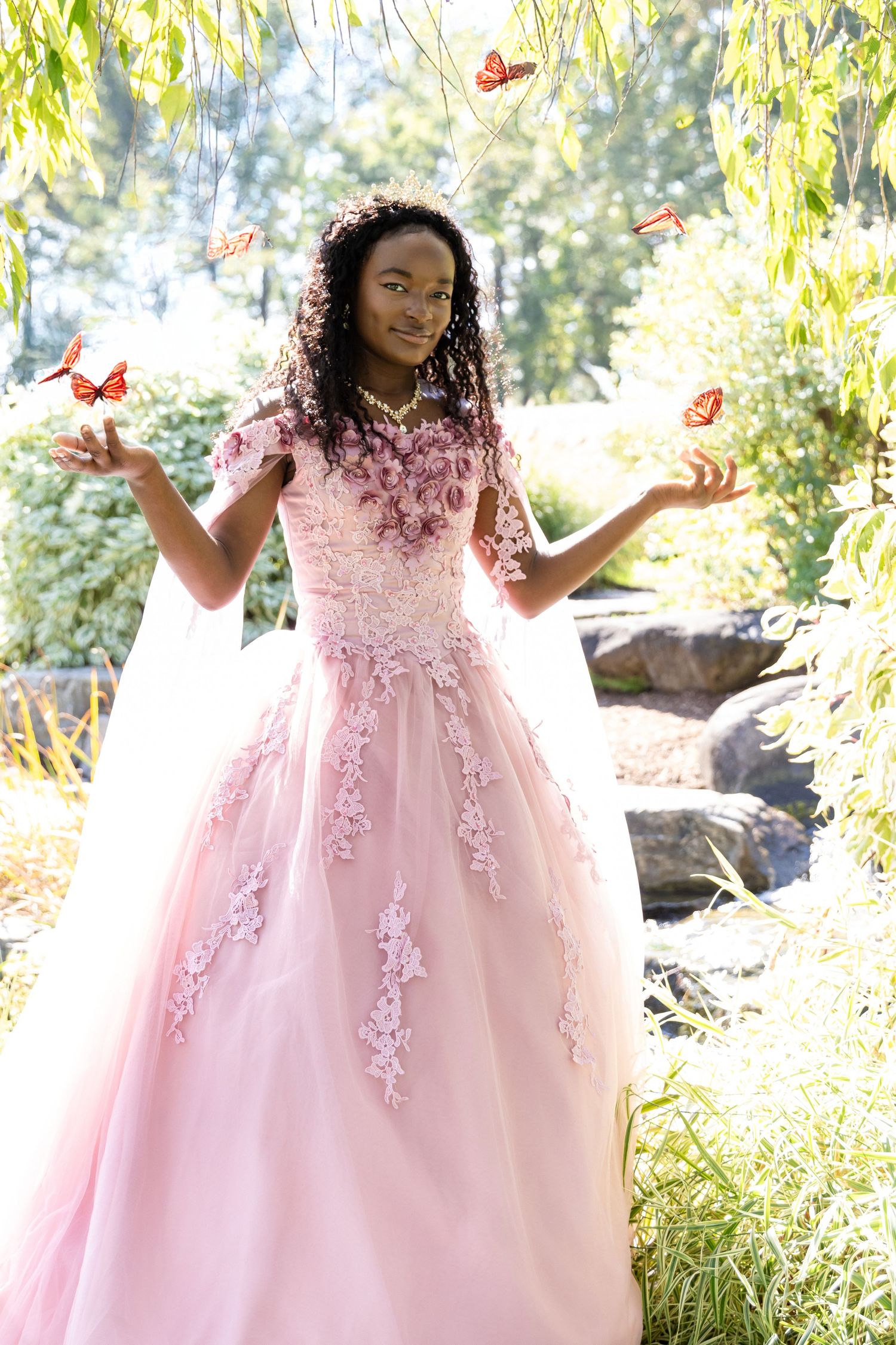 Young woman in a pink tulle gown reaches out her hands as orange butterflies flutter around her in a leafy garden.