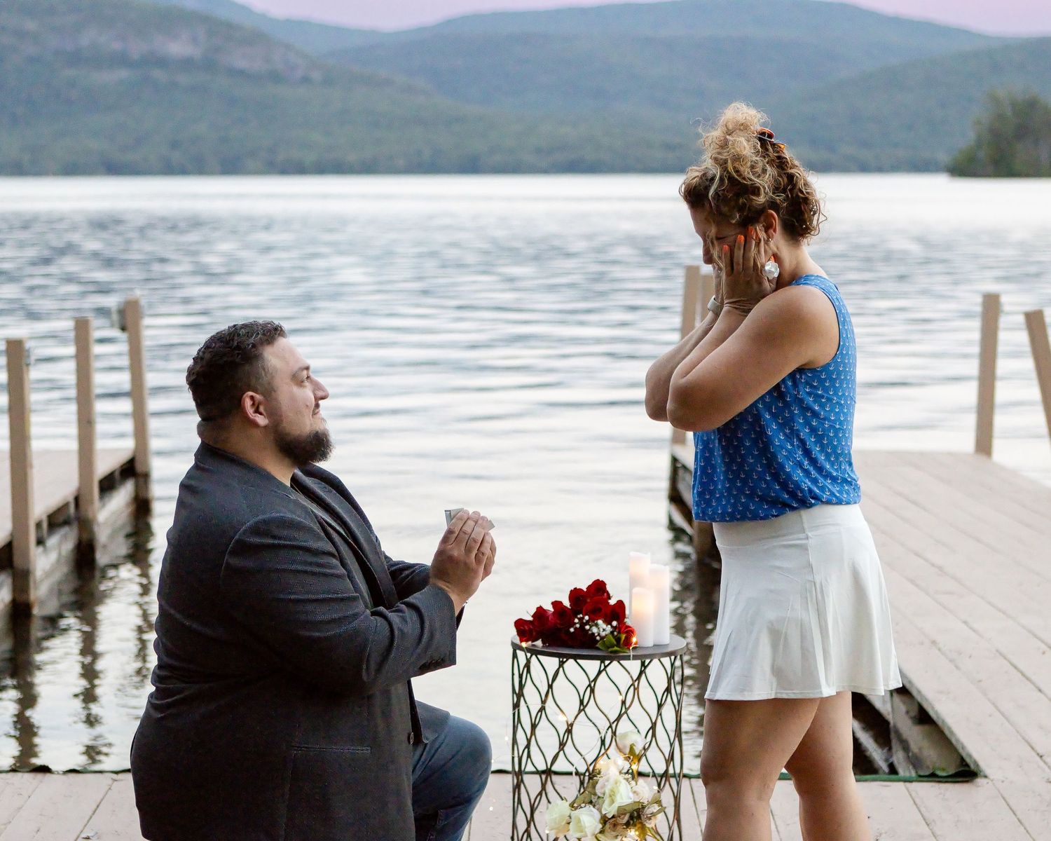 A man kneels on one knee proposing to a woman in a blue top and white skirt on a lakeside dock with roses and candles.