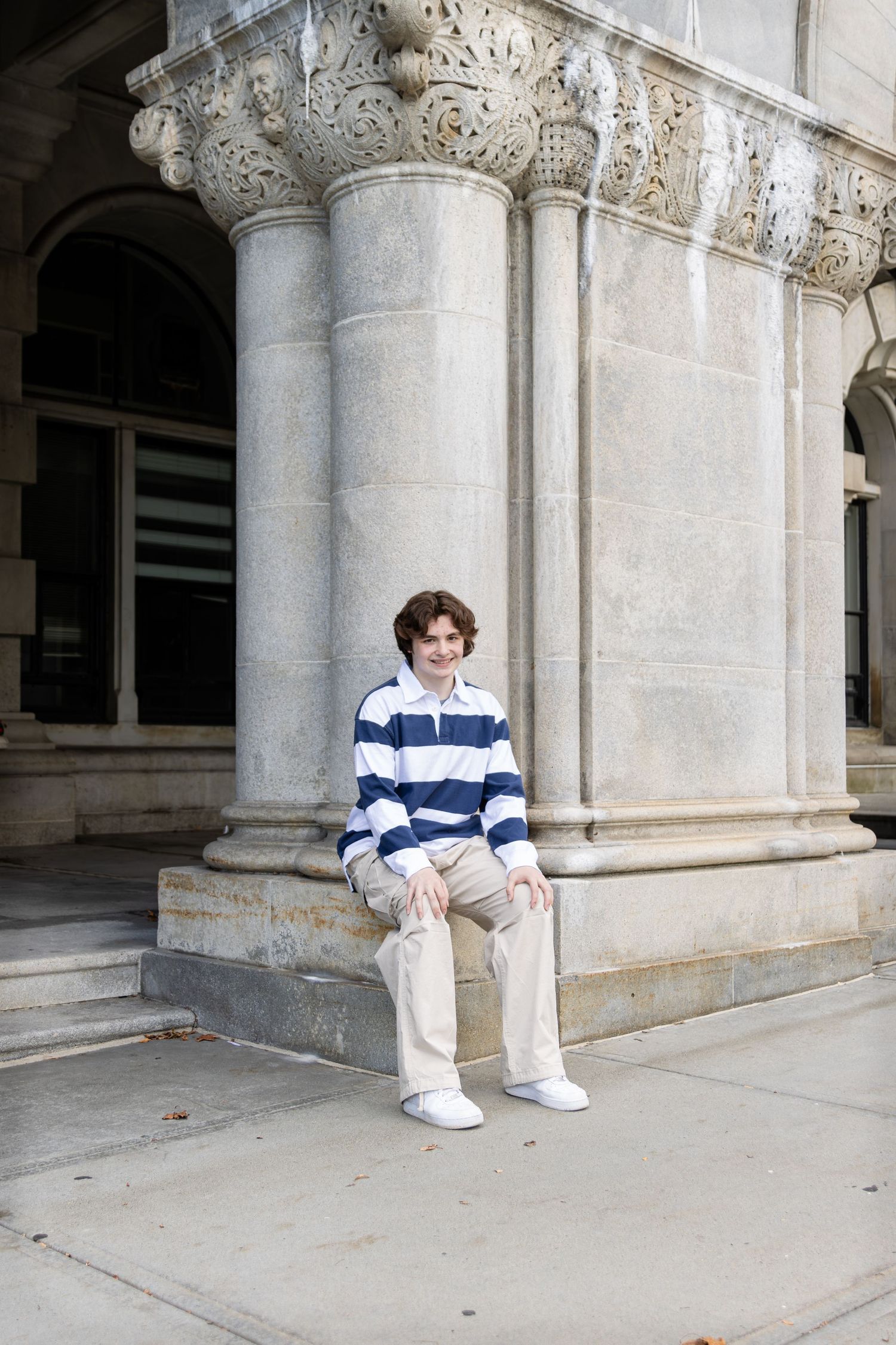 Teenager in a blue and white striped shirt and khaki pants sits on the base of a stone column with ornate carvings.