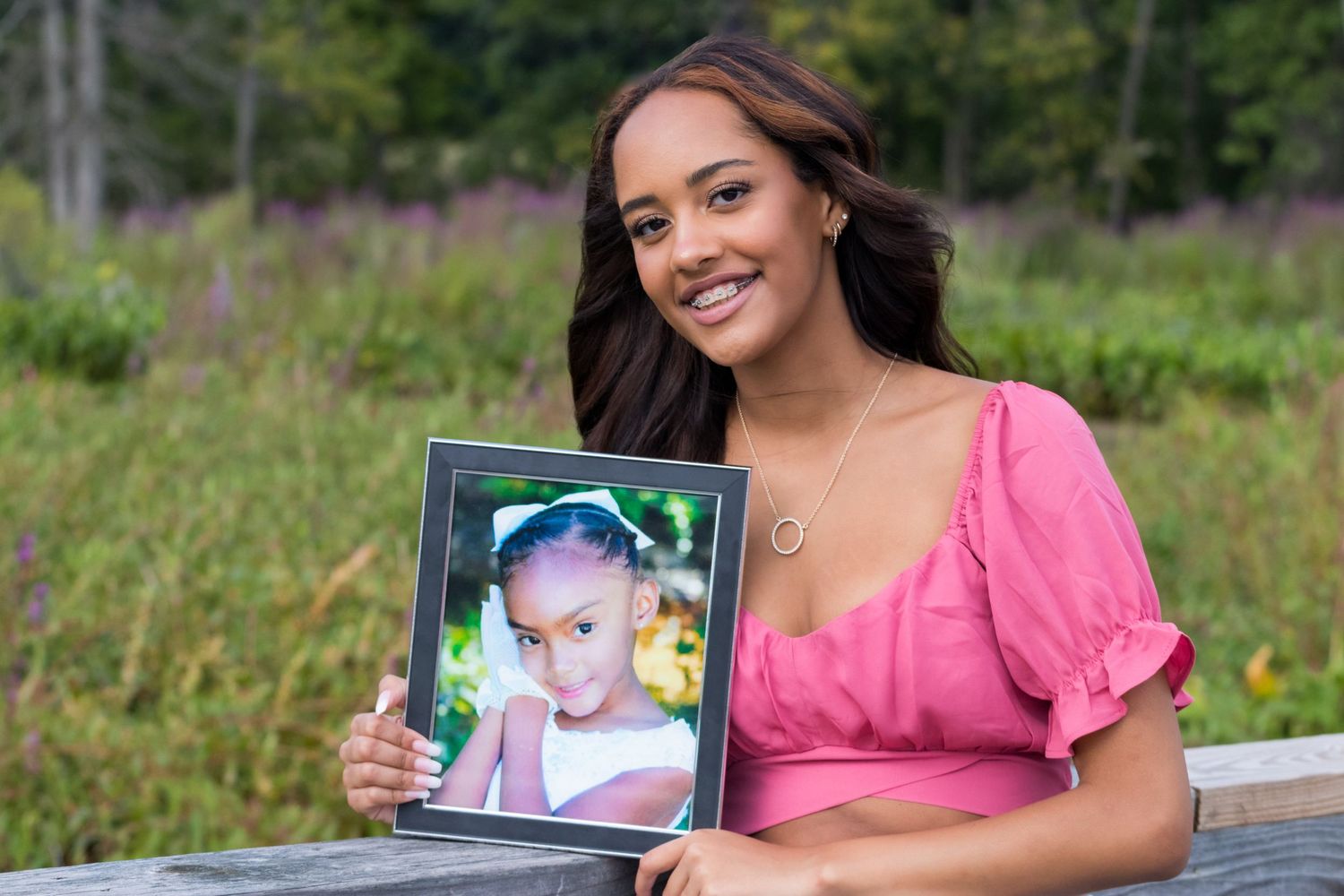 Smiling young woman in a pink puff-sleeve top holds a framed childhood portrait while standing in a green field.