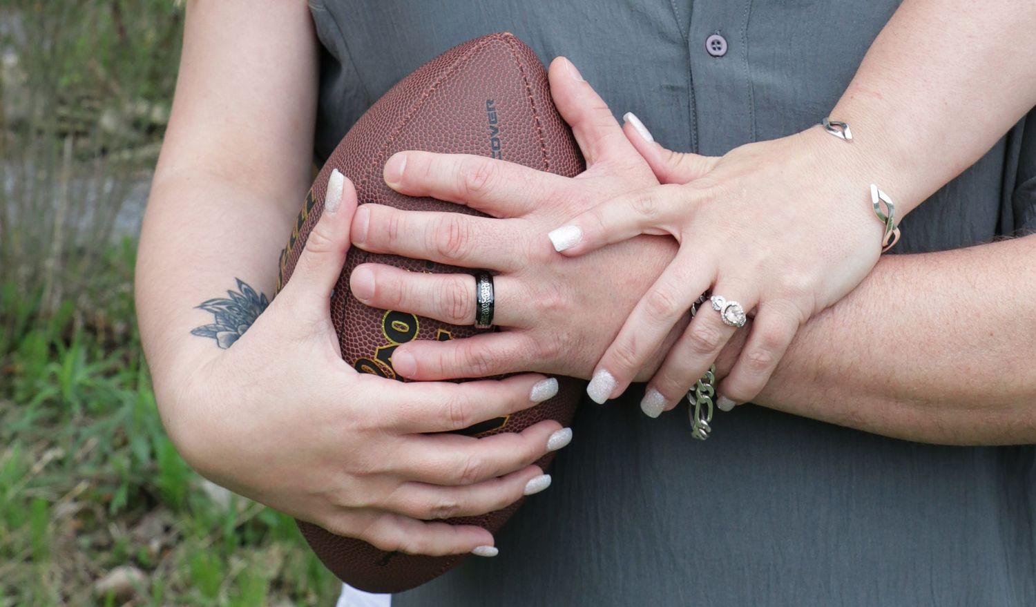 A couple holds a football while showing their wedding rings and her engagement ring, with a floral tattoo on her arm.