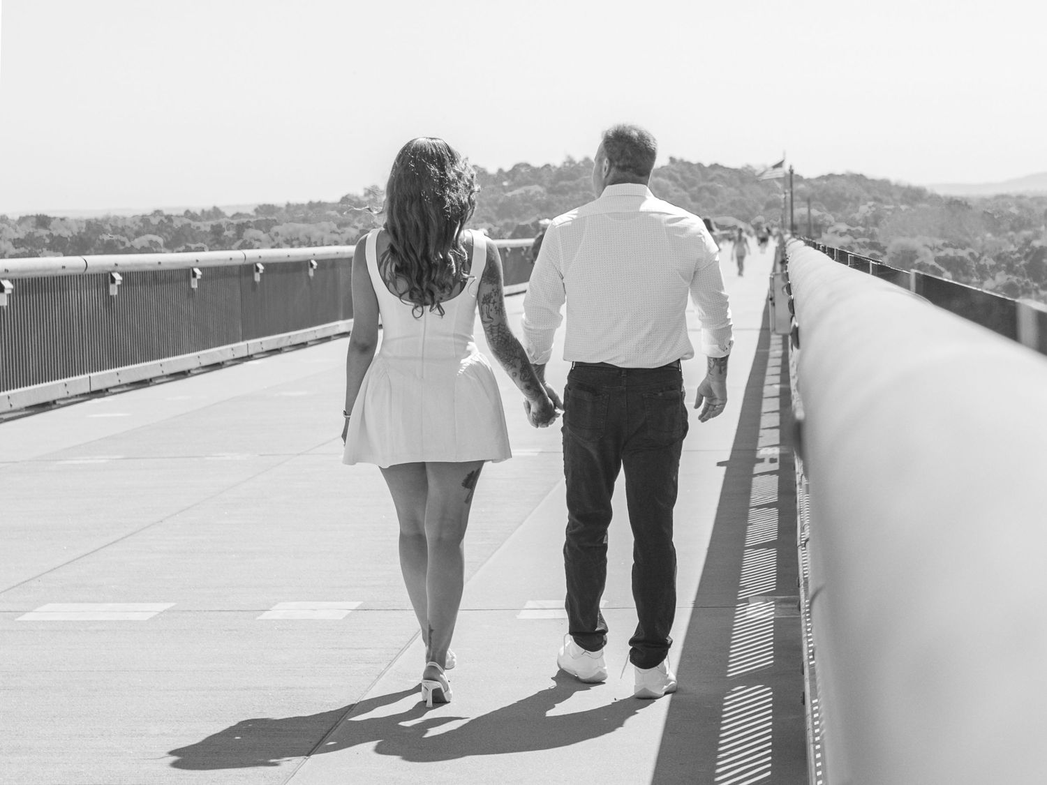 Black and white photo of a couple holding hands and walking away along a bridge walkway with hills in the distance.