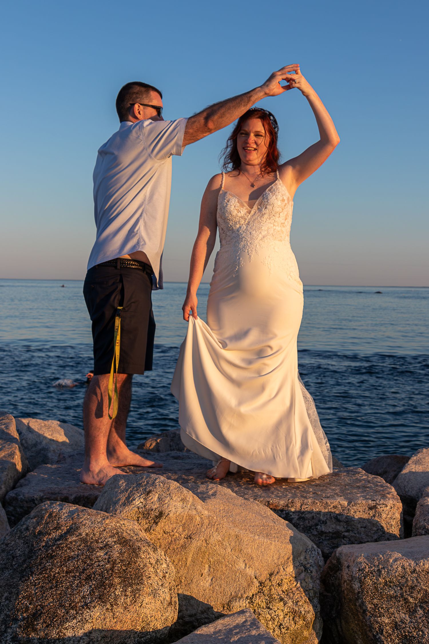Couple dances on coastal boulders at sunset as the bride in a white lace gown lifts her skirt while the groom twirls her by the ocean.