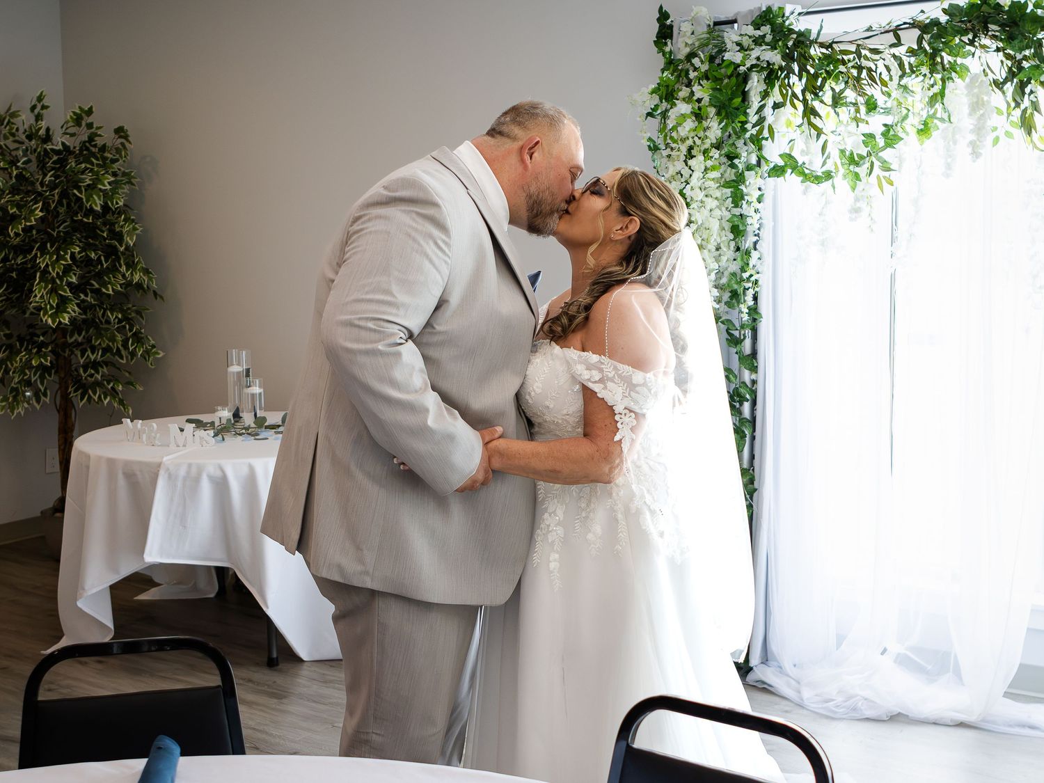 A groom in a gray suit kisses his bride in a white lace gown and veil beside a decorated table in a bright wedding venue.