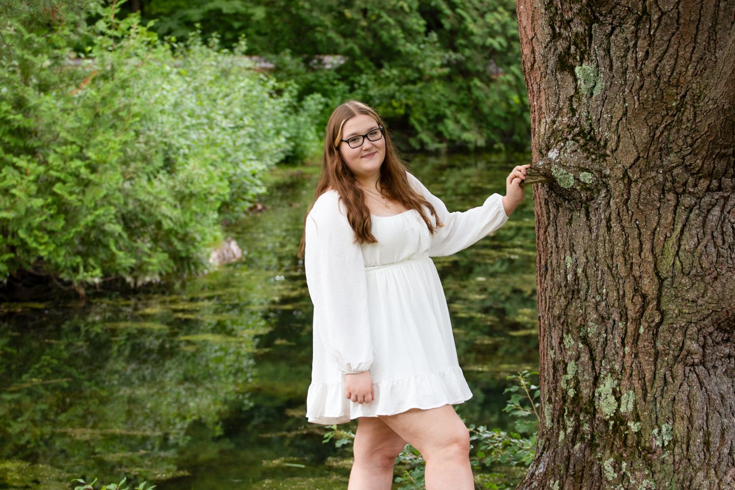 Young woman with glasses in a white dress poses against a large tree trunk beside a calm, green pond.
