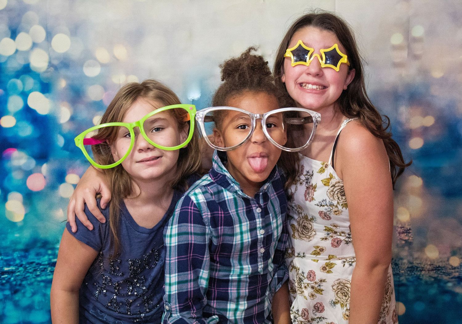 Three smiling girls pose together in oversized novelty glasses, one sticking out her tongue, against a sparkling blue bokeh party backdrop.