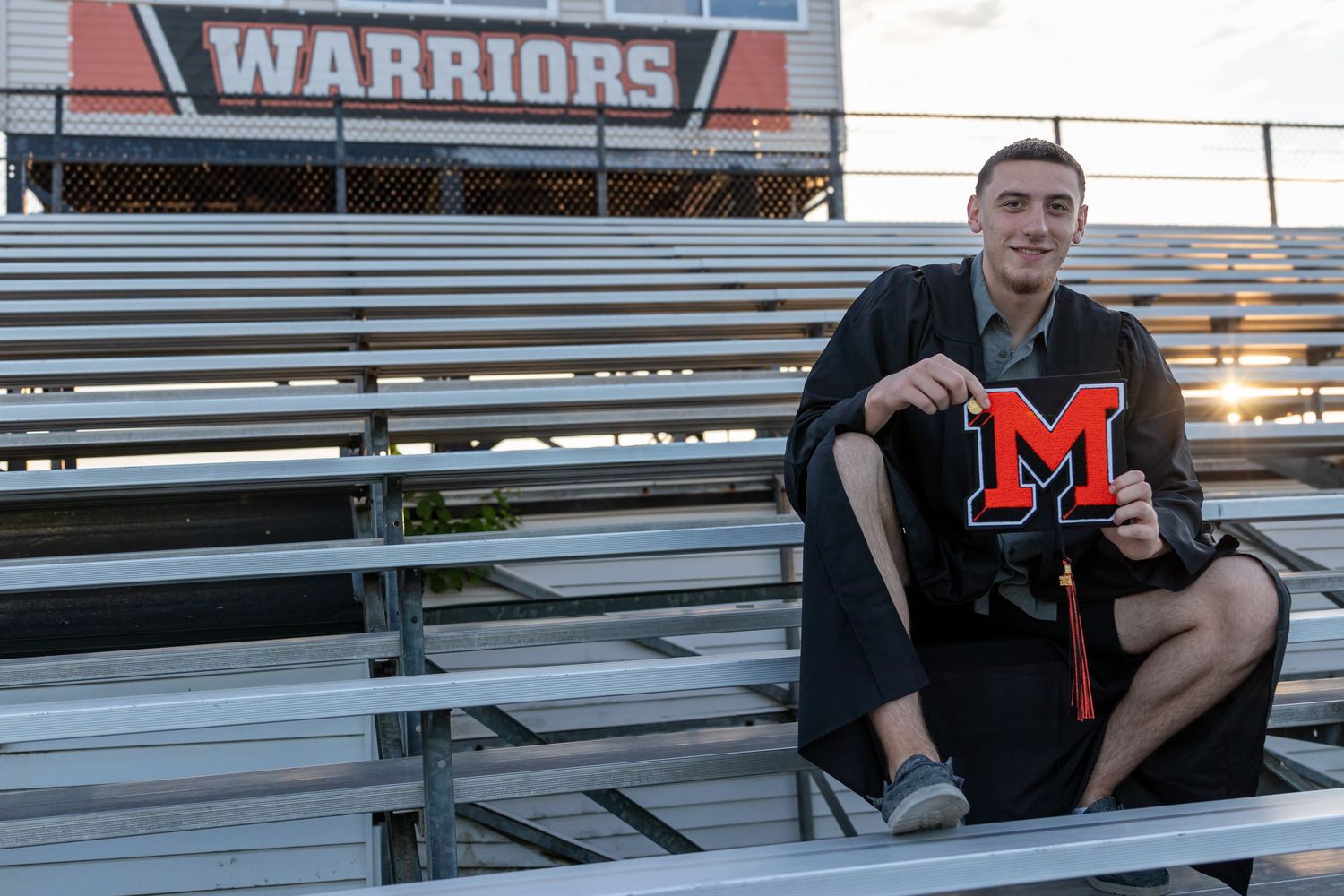 A smiling graduate in a black gown sits on empty bleachers beneath a Warriors banner, holding a mortarboard with a red block M.