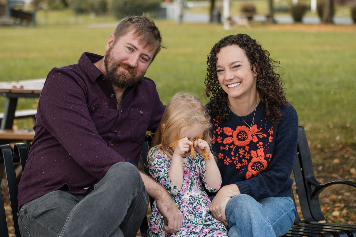 A smiling bearded man and a curly-haired woman sit on a park bench with a young girl wiping her eyes between them.
