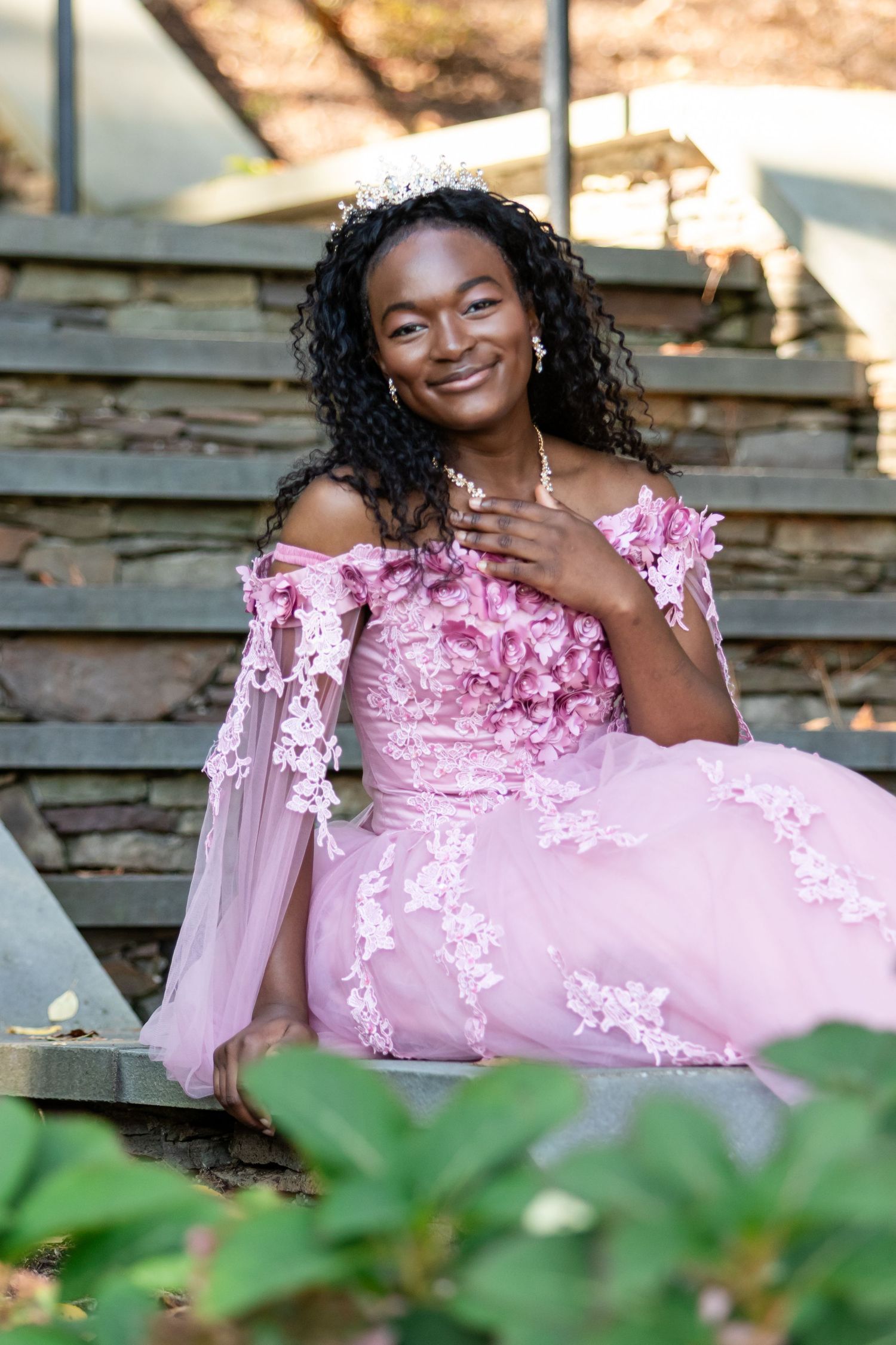 Smiling young woman in a pink off-the-shoulder floral ball gown and tiara sits on stone steps outdoors.