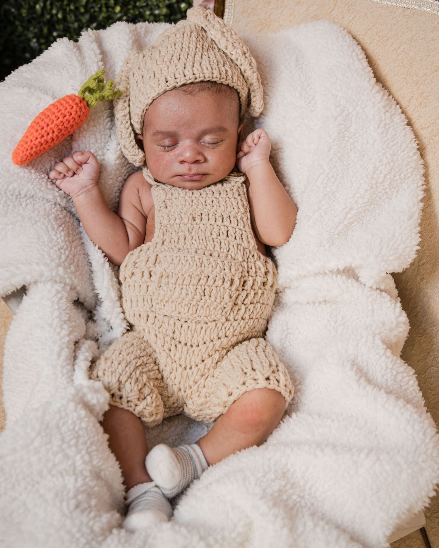 A sleeping newborn baby wearing a knitted bunny outfit rests on a fluffy white blanket next to a crocheted carrot.
