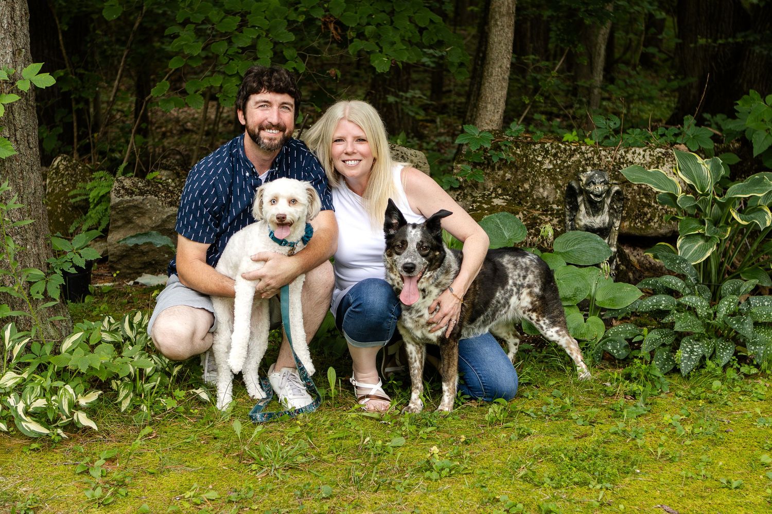 A smiling man and woman kneel on grass with their white dog and a blue heeler in a leafy garden.