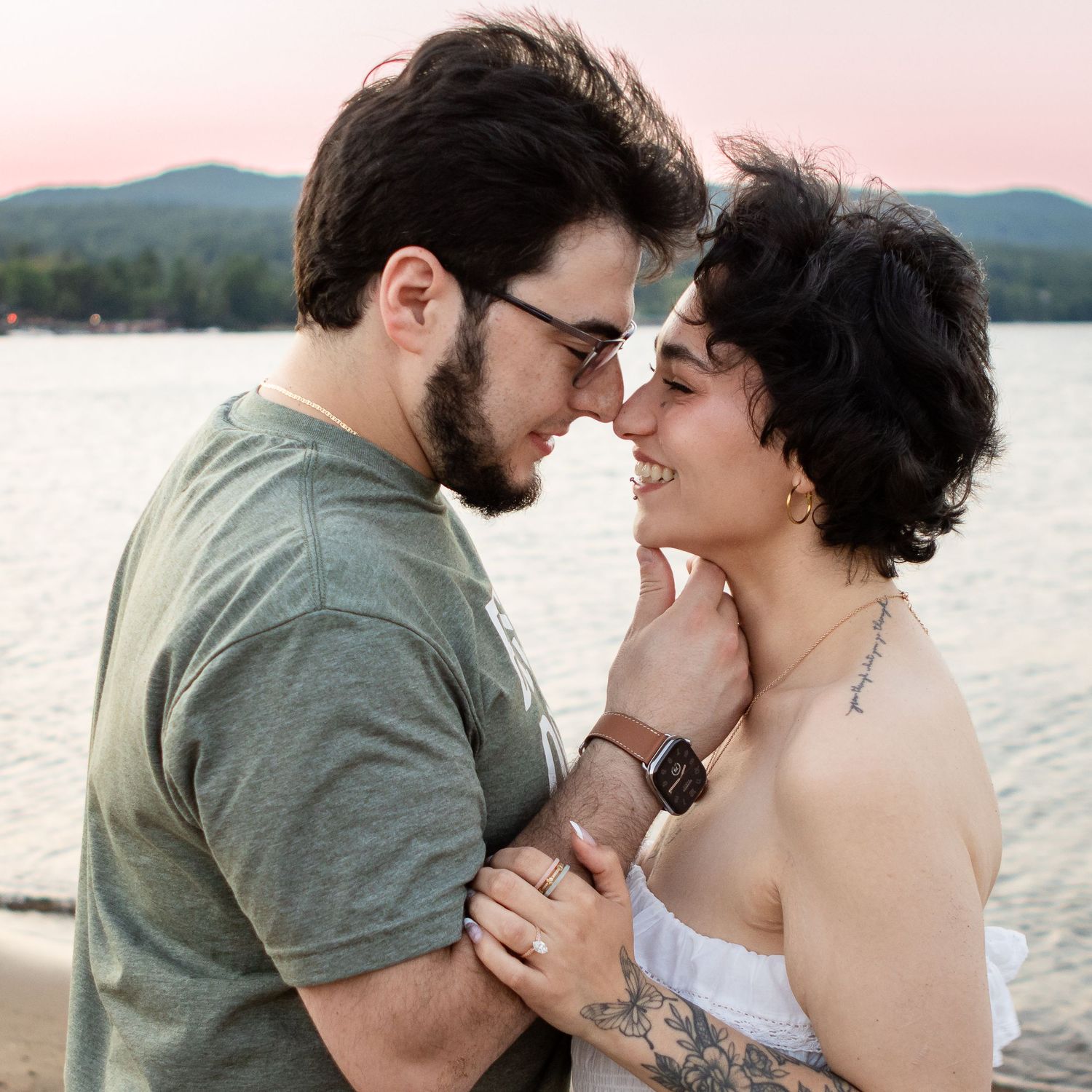 Couple touching foreheads and smiling by a lake at dusk, with hills and a pink sky behind them.