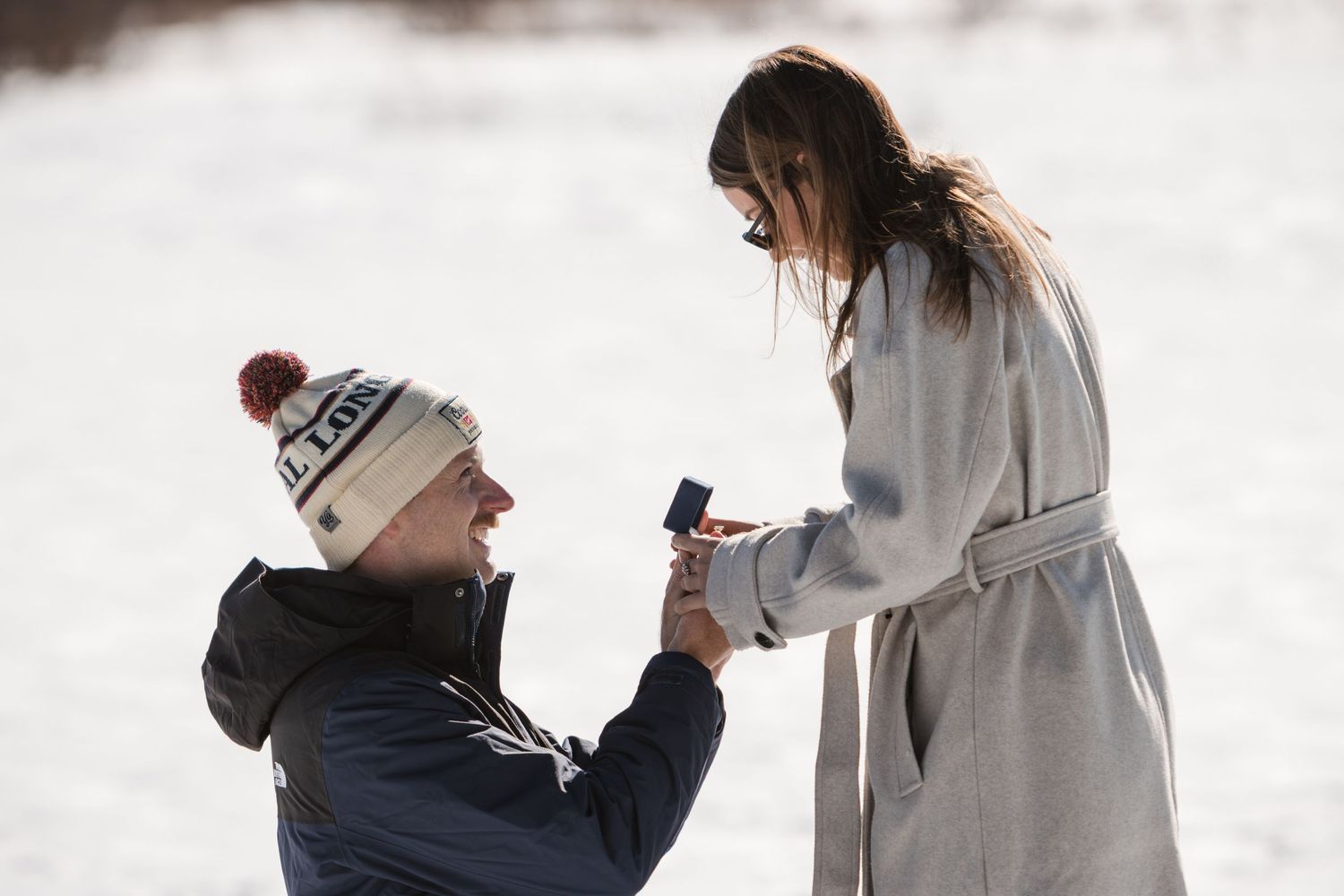 A man in a beanie kneels in the snow holding a ring box as he proposes to a woman in a long gray coat.