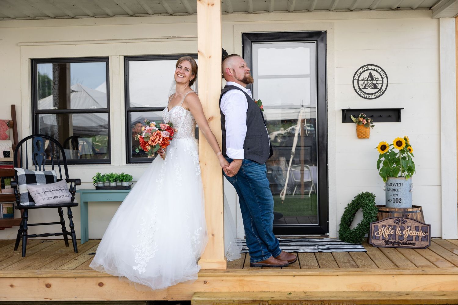 A bride in a lace wedding gown holds a bouquet and the hand of her groom on a rustic wooden porch.