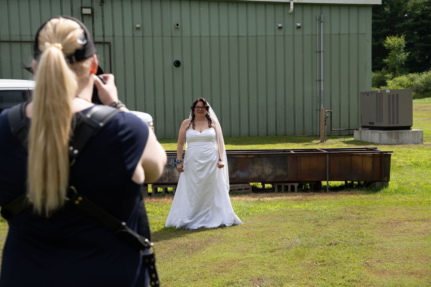 Photographer with a blonde ponytail takes a photo of a bride in a white gown and veil standing on grass near a green building.