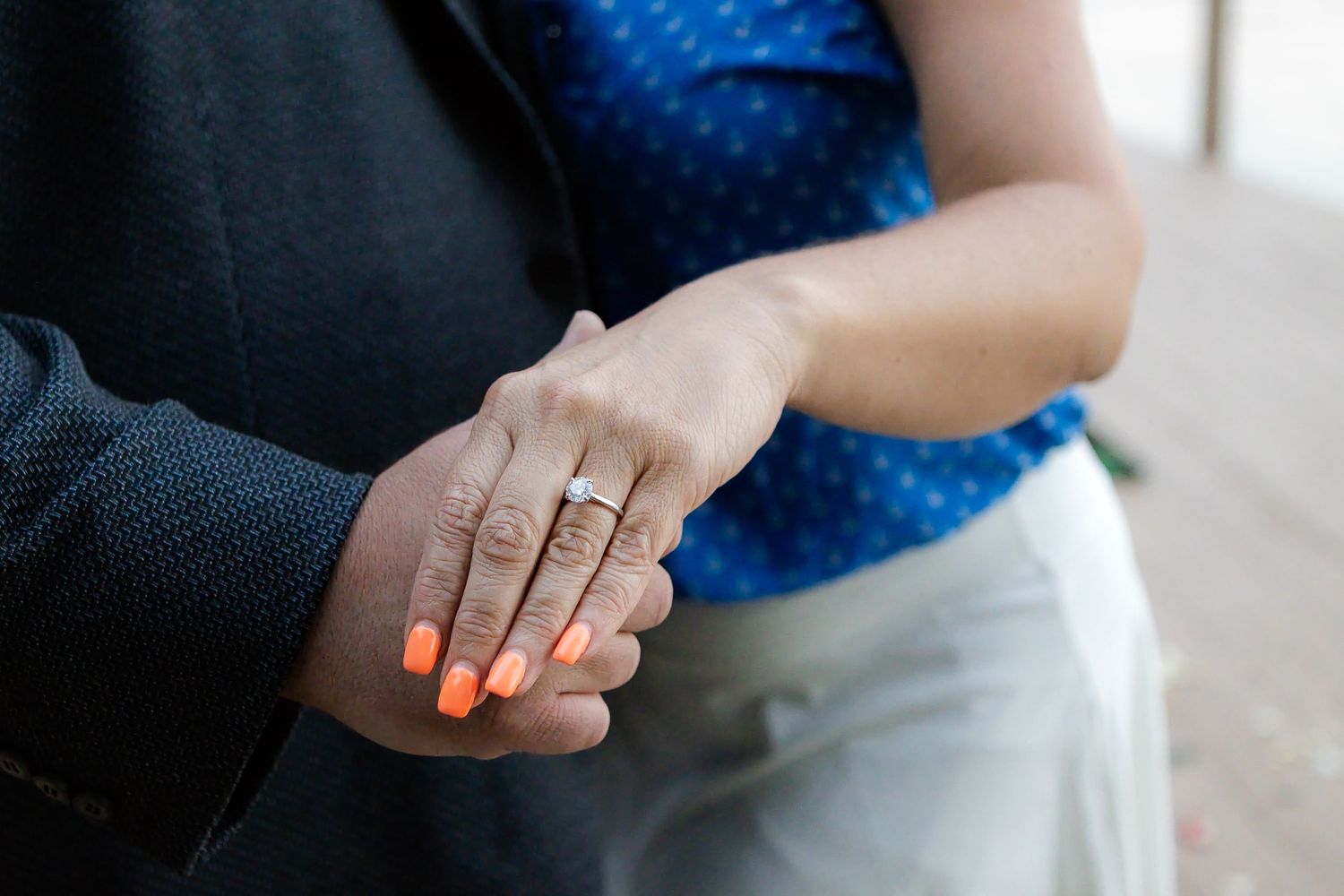 Close-up of a couple's hands showing a woman's sparkling engagement ring and bright orange manicured nails.