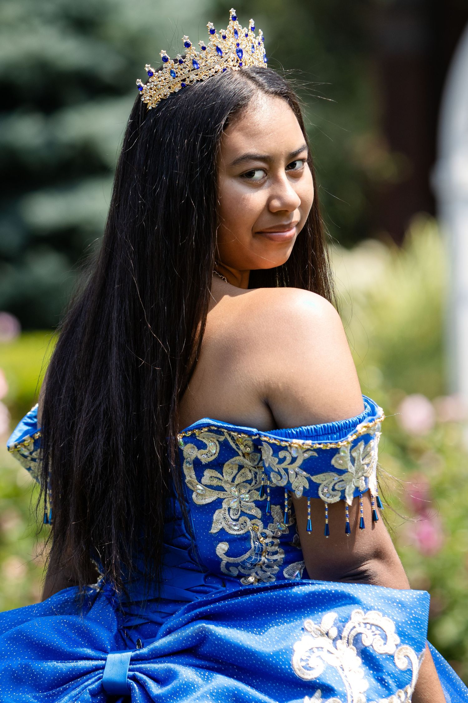Young woman in a blue embroidered quinceañera gown and gold tiara looks over her shoulder in a sunny garden.