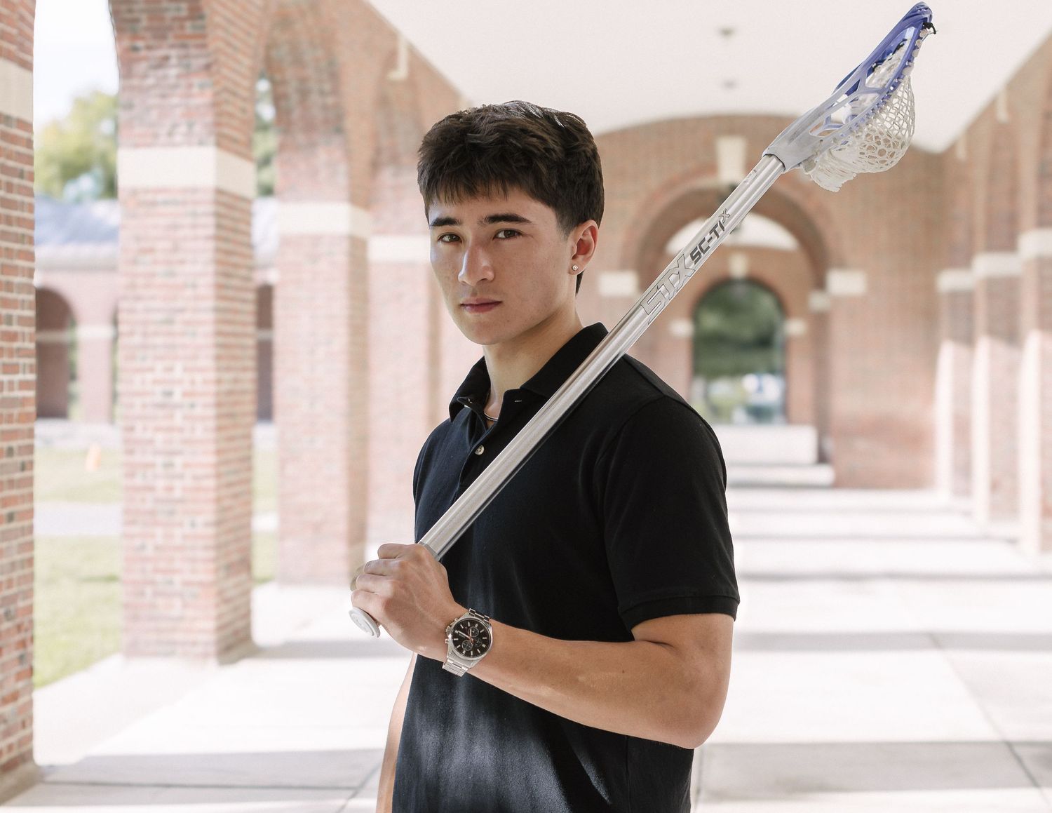 Young man in a black polo holds a lacrosse stick across his body while standing in a brick-columned outdoor corridor.