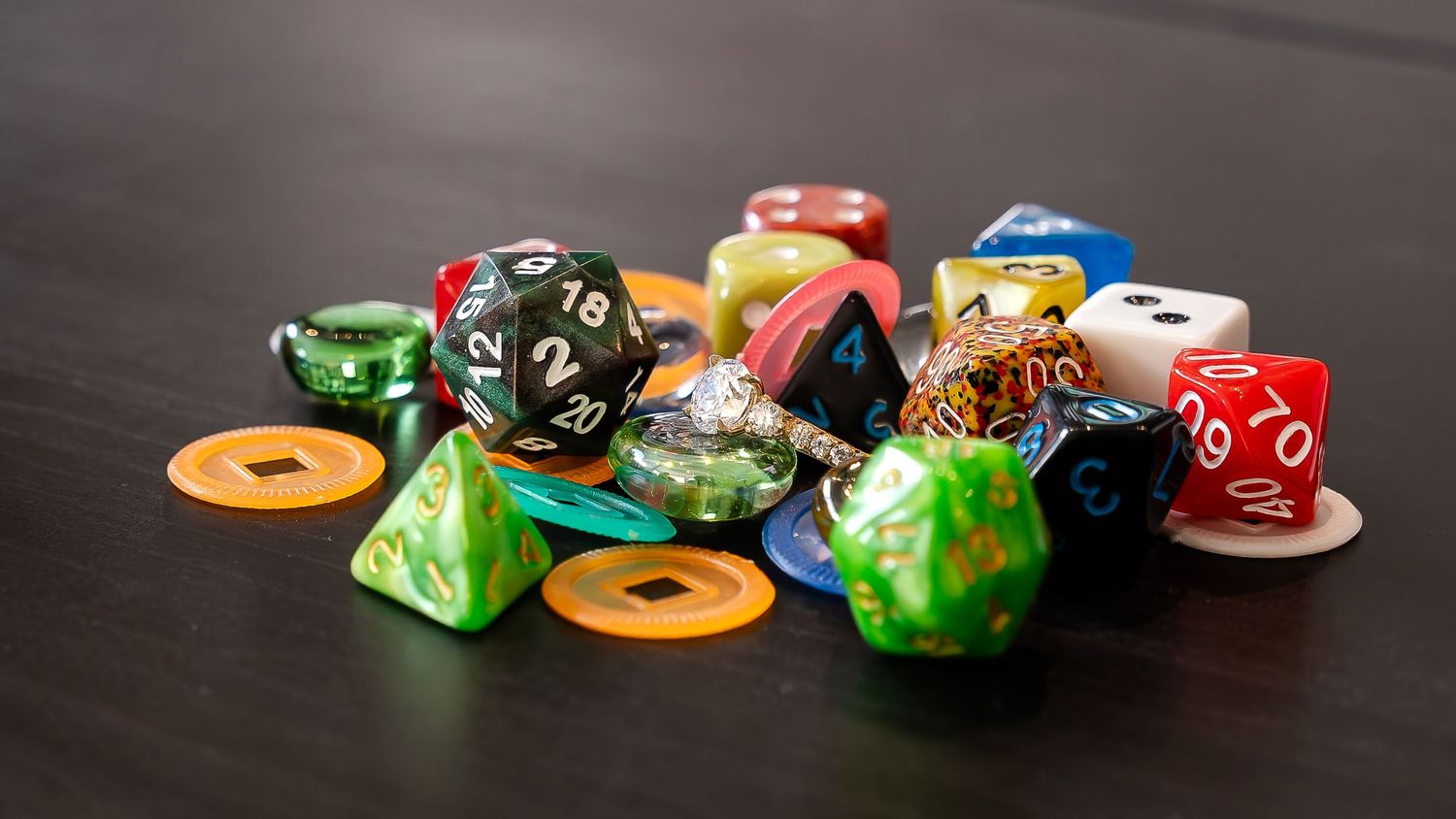 Pile of colorful polyhedral tabletop gaming dice and orange tokens on a dark wooden table.