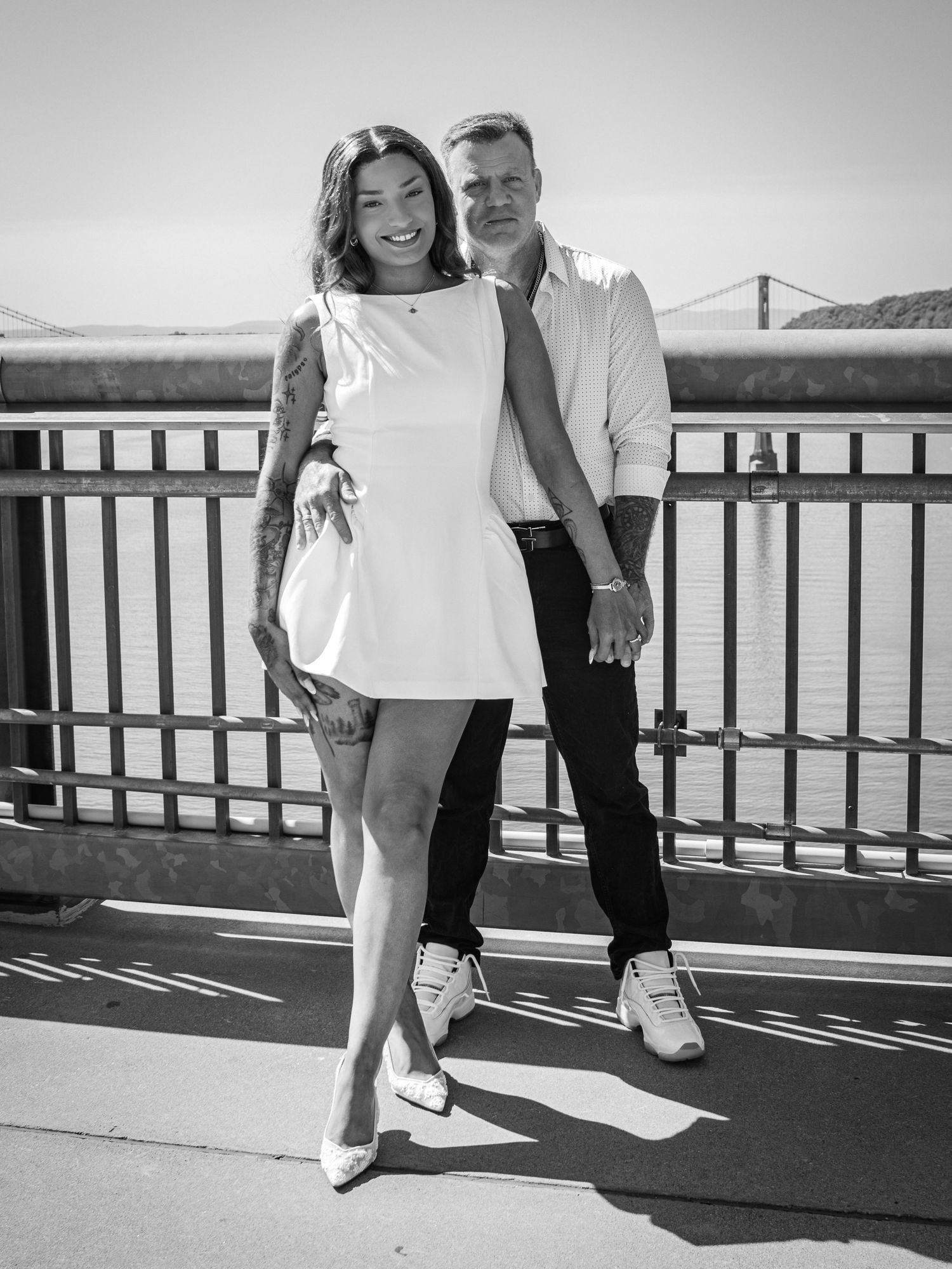 Black and white portrait of a smiling couple in white outfits leaning against a bridge railing with water and a bridge behind.