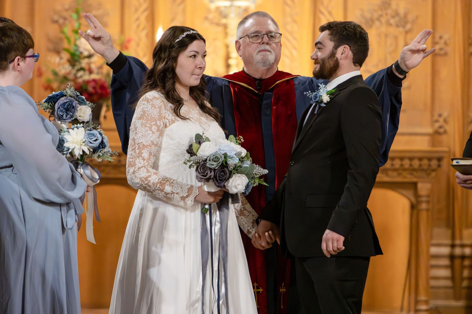 Bride and groom hold hands at the altar as an officiant in a red and navy robe raises a hand in blessing during a church ceremony.