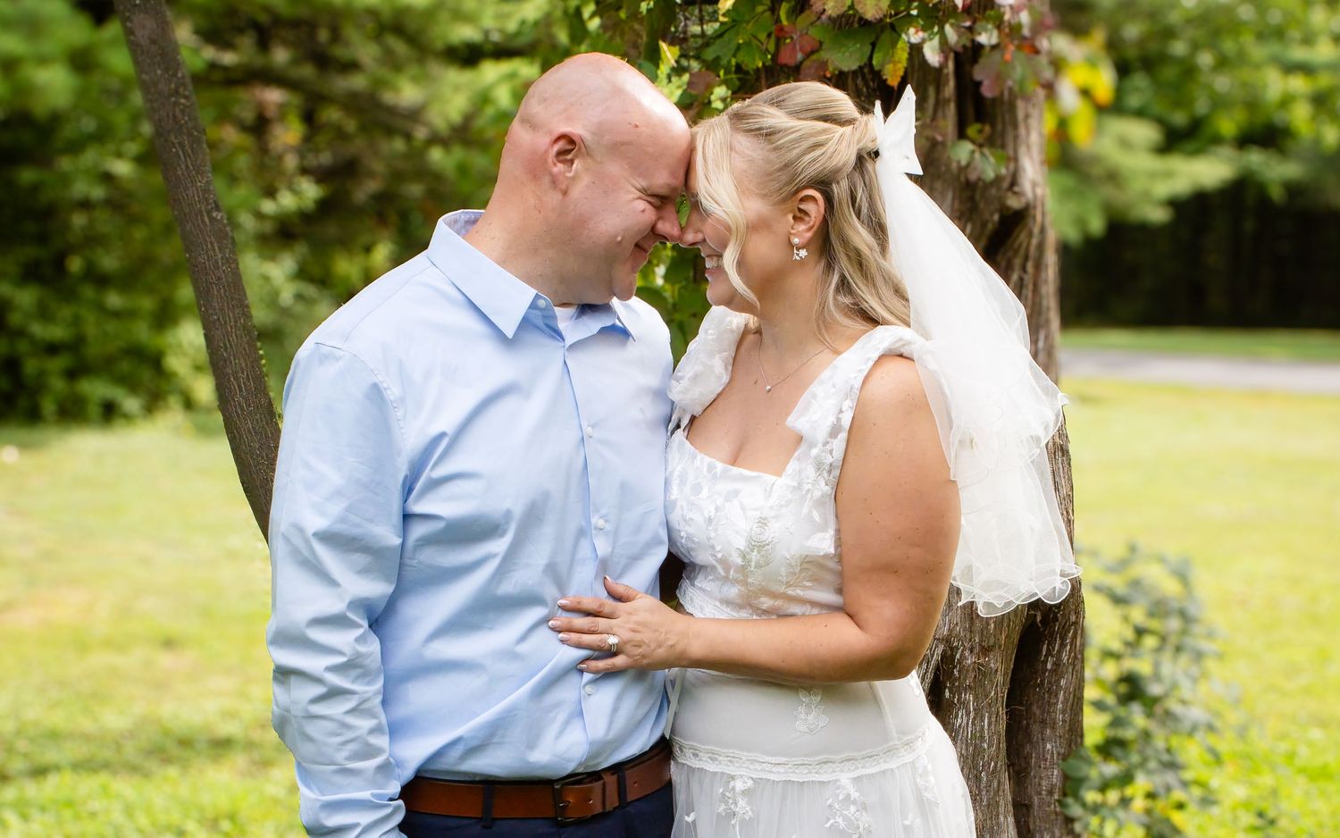 A smiling bride in a white lace dress and veil and a groom in a light blue shirt touch foreheads outdoors on a grassy lawn.