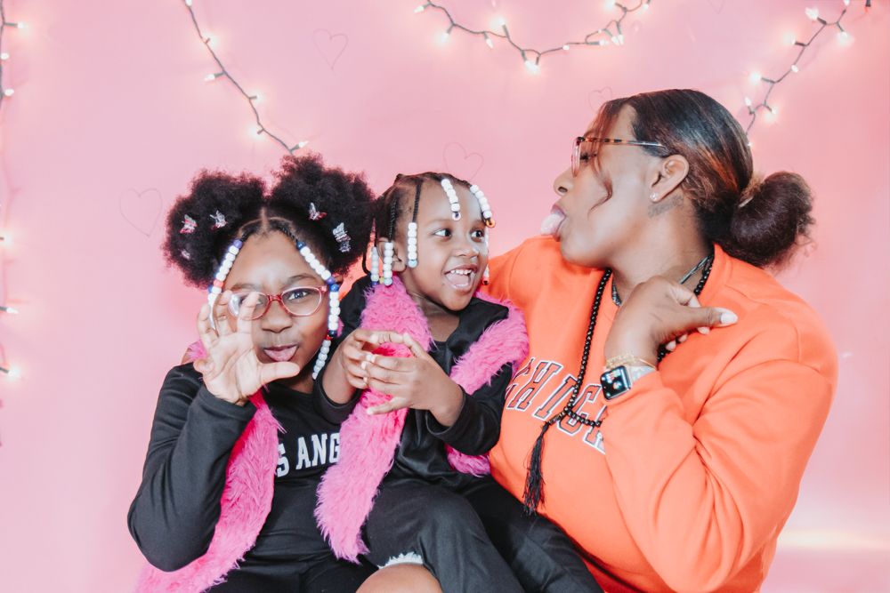Mother in an orange sweatshirt poses with two young girls in pink feather scarves against a pink backdrop with string lights.