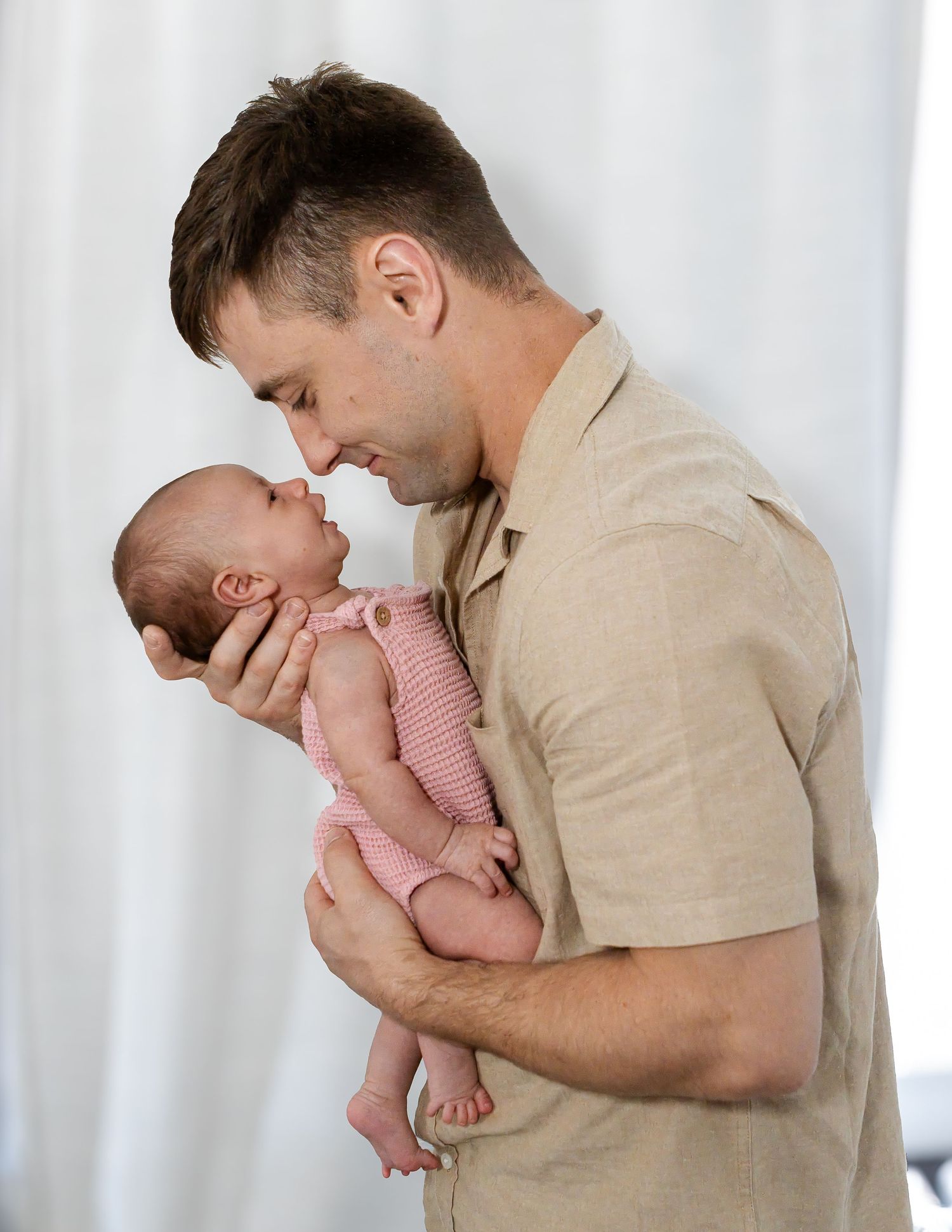 A father in a beige shirt gently holds his newborn baby in a pink knit outfit, touching noses in front of white curtains.