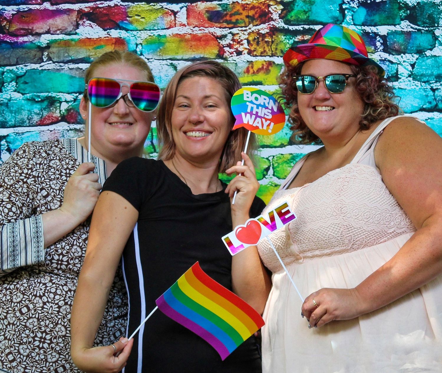 Three smiling women pose in a photo booth with rainbow sunglasses, a pride flag and fun signs in front of a colorful graffiti brick wall.