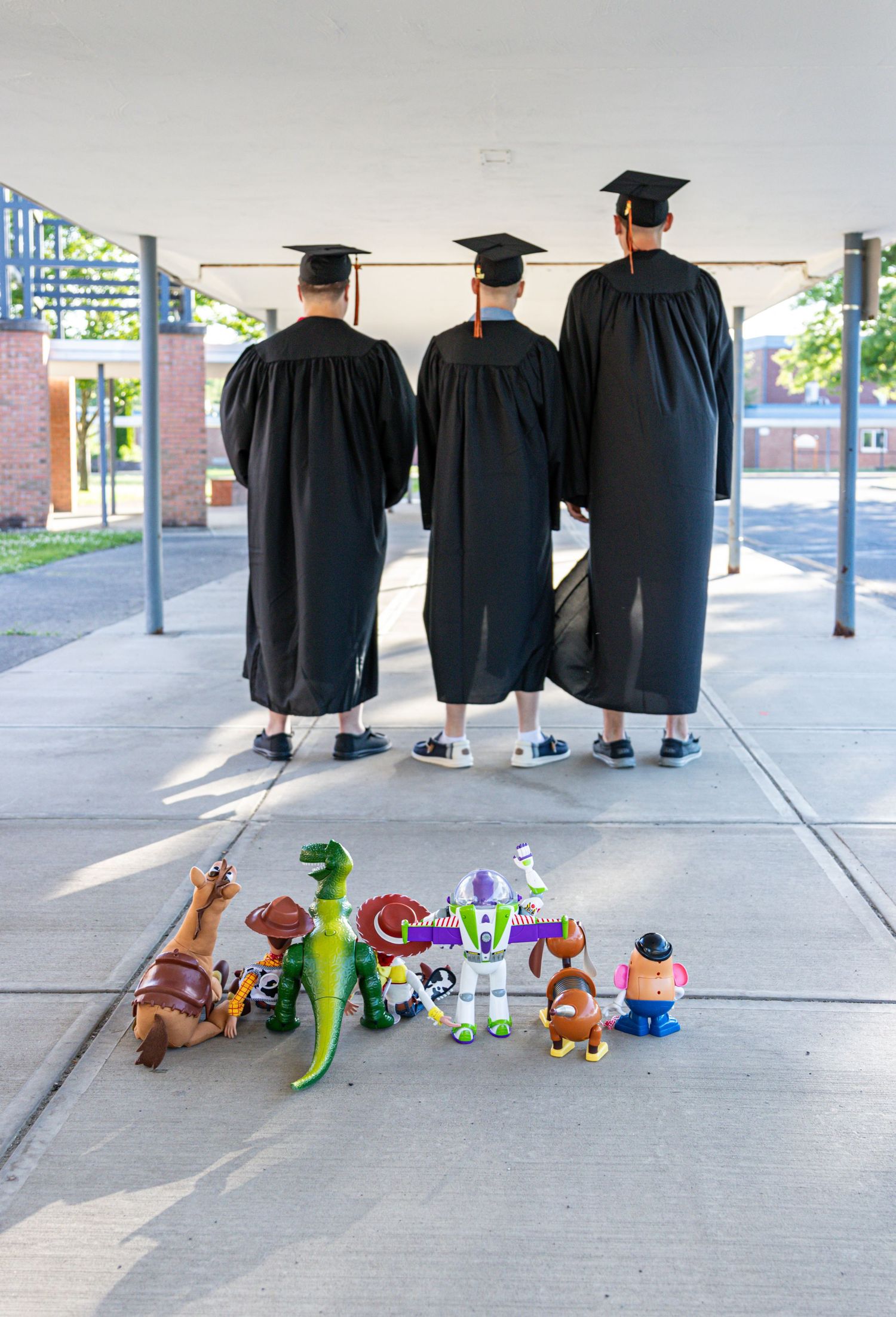 Three graduates in caps and gowns face away from the camera toward a lineup of Toy Story figures including Buzz Lightyear and Rex.