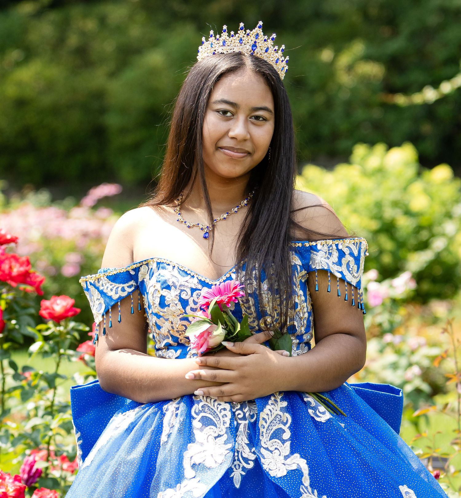 Young woman in a blue off-shoulder quinceañera dress and tiara holds pink roses in front of a blooming garden.
