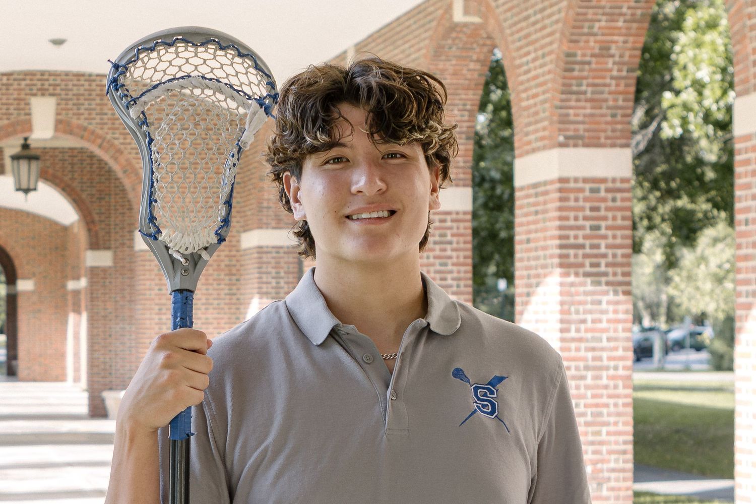 Smiling young man in a gray polo holds a lacrosse stick while standing in front of red brick arches.