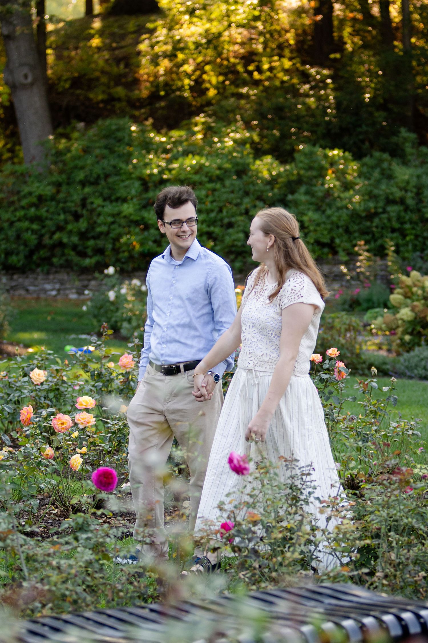 A smiling couple holds hands and walks through a blooming rose garden during an outdoor engagement photo session.
