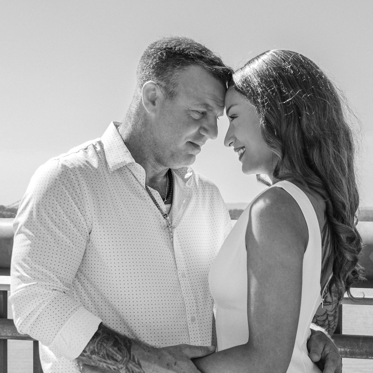 Black and white close-up of a tattooed man and a long-haired woman touching foreheads in a tender embrace outdoors.