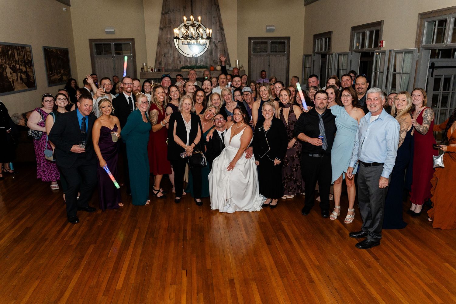 Large group of wedding guests in formal attire gathers around the bride and groom for a group photo in a elegant hall with wood floors.