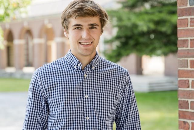 Smiling young man in a blue checkered button-up shirt stands outdoors near a brick building and green trees.