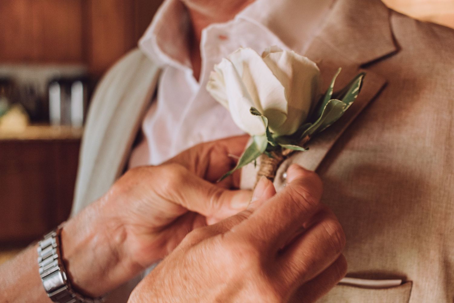 Hands pin a white rose boutonniere onto a suit jacket lapel before a wedding.